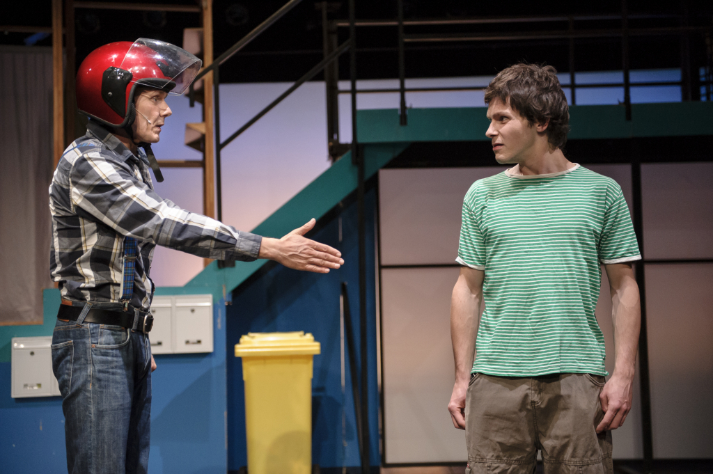 Zwei Schauspieler, einer in einem Motorradhelm, interagieren auf der Bühne. Der Schauspieler mit Motorradheld reicht dem anderen die Hand.
