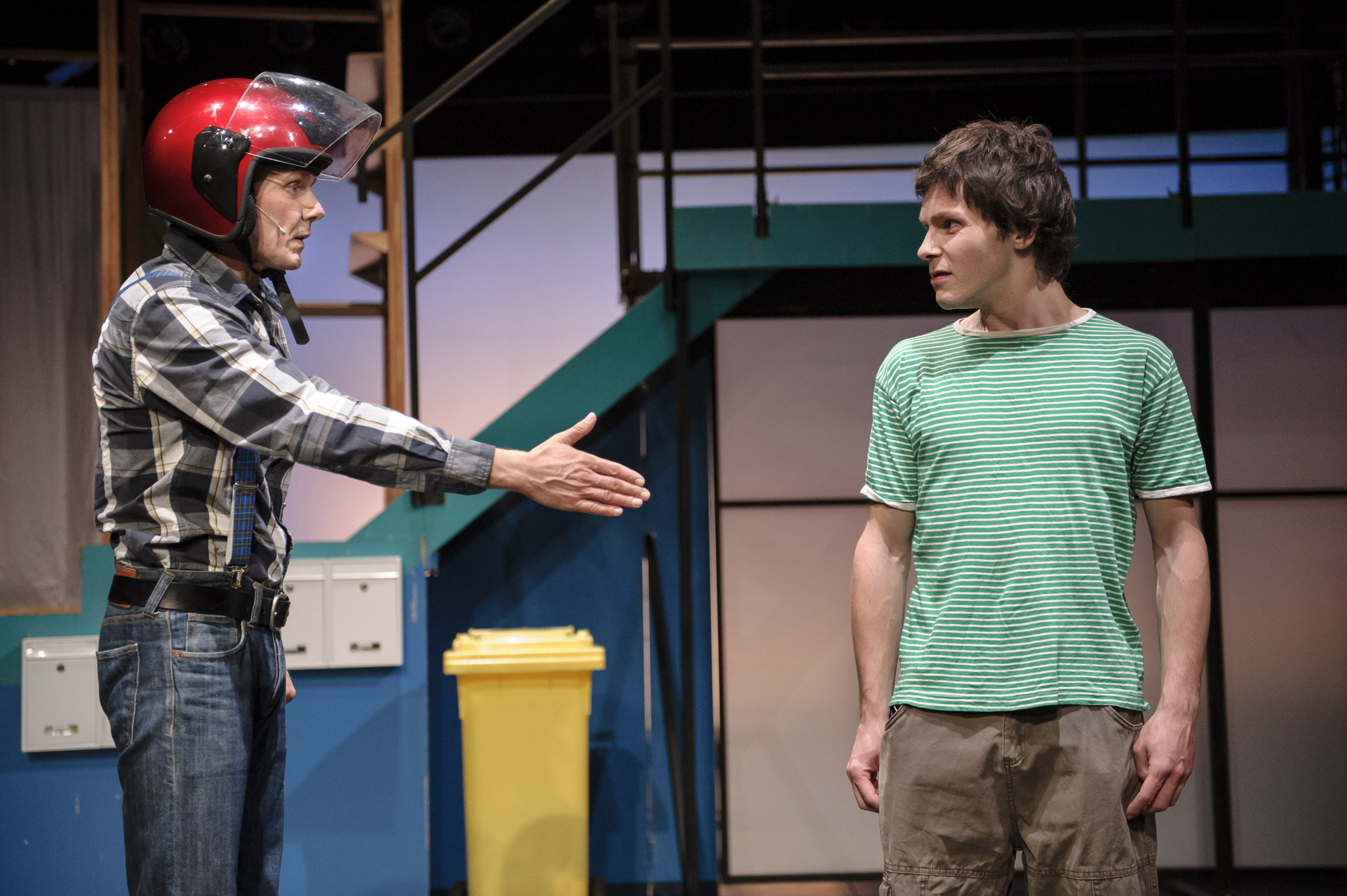 Zwei Schauspieler, einer in einem Motorradhelm, interagieren auf der Bühne. Der Schauspieler mit Motorradheld reicht dem anderen die Hand.