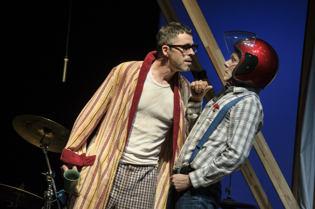 Zwei Schauspieler auf der Bühne: einer im Bademantel mit Kaffeetasse, der andere mit Helm.