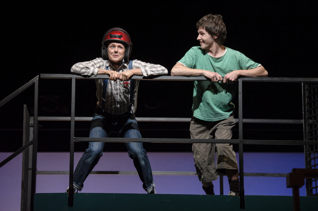 Zwei Schauspielern lehnen auf einem Geländer, einer trägt einen Helm.