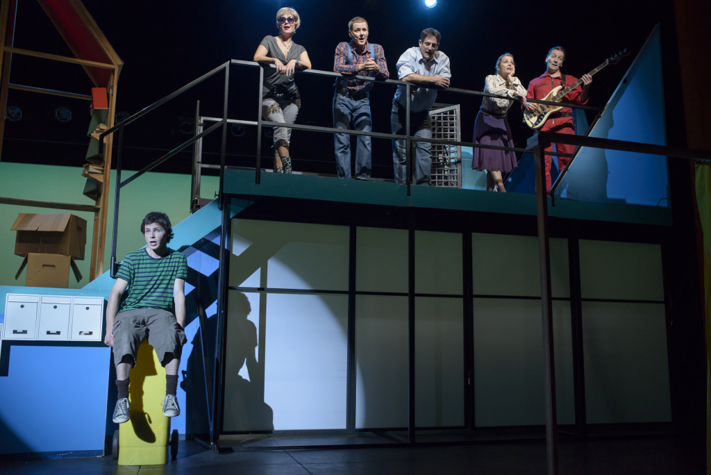 Fünf Schauspieler*innen stehen auf einem Balkon und schauen auf eine Schauspieler, der unten sitzt.