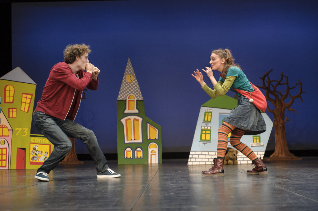 Zwei Personen in bunter Kleidung stehen sich auf einer Bühne mit bunten Häuserkulissen gegenüber, beide in spielerischer Pose.