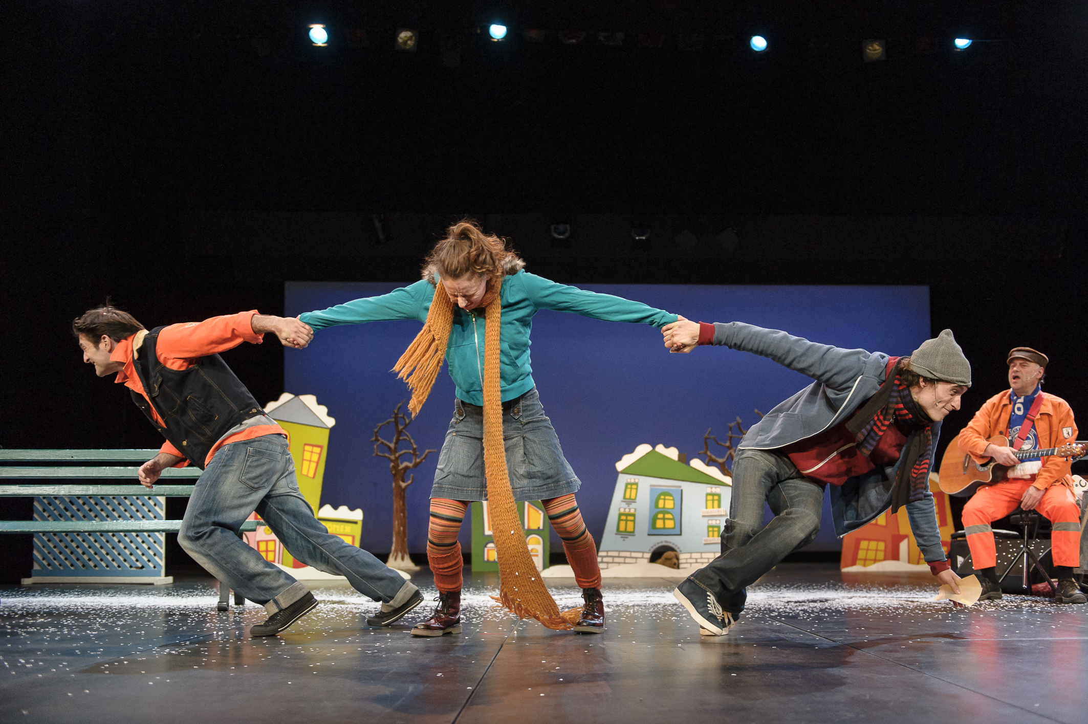 Drei Personen auf Bühne halten sich an den Händen, lehnen sich nach außen, bunte Kleidung, im Hintergrund Bühnenbild mit Häusern und Bäumen.