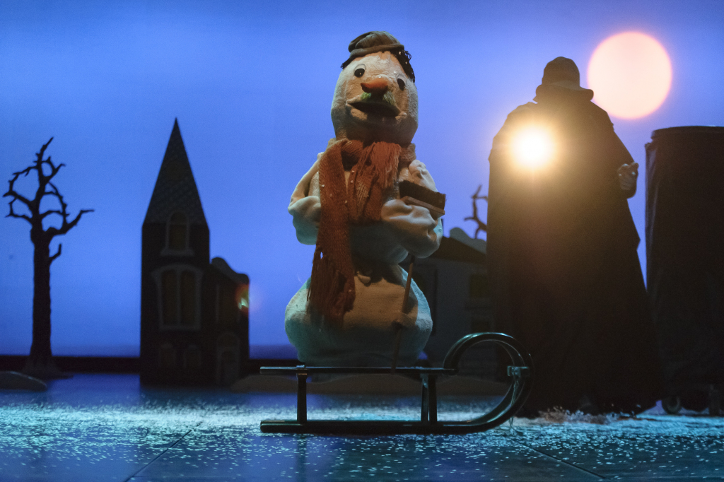 Schneemann mit braunem Schal und Mütze auf einem Schlitten, im Hintergrund ein kahler Baum, ein Kirchturm und eine dunkle Figur vor einem großen Lichtpunkt.