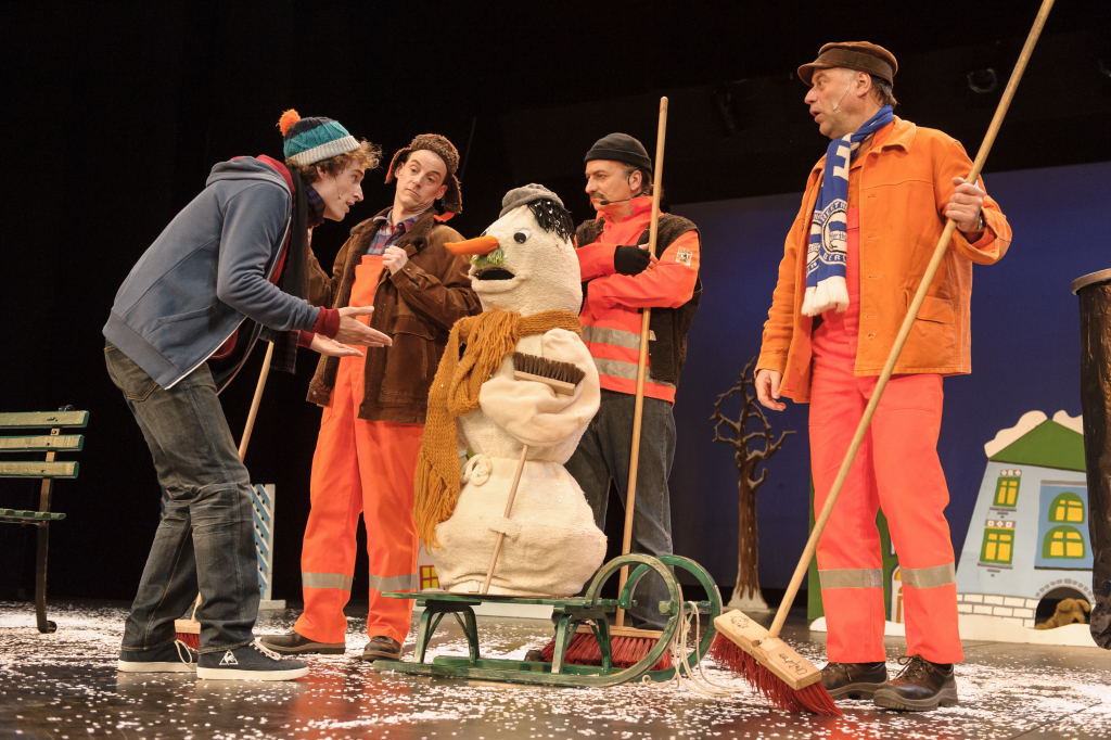Vier Personen in oranger Arbeitskleidung und eine Person in blauer Jacke stehen um eine Schneemannfigur mit orangefarbenem Schal und schwarzer Mütze auf einem Schlitten.