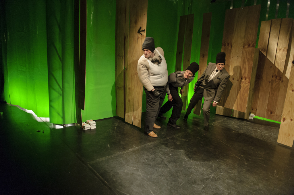 Szenenbild mit drei Schauspielern in Anzügen und Mützen auf einer Bühne vor grüner Kulisse.