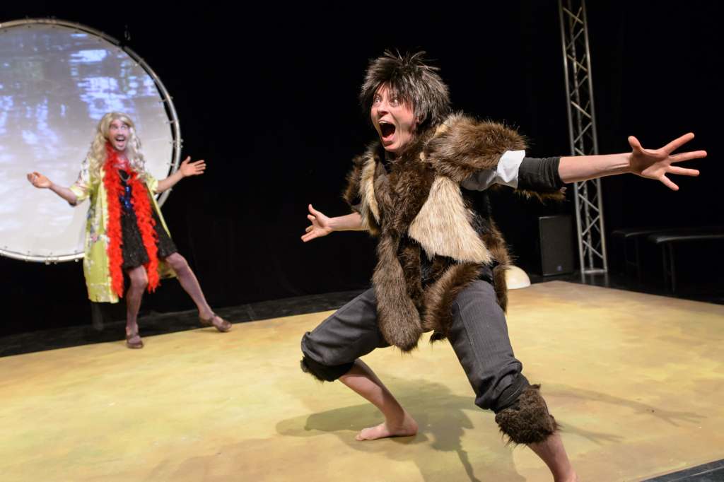 Person in braunem Fellkostüm mit ausgestreckten Armen und gebeugten Knien auf Bühne, im Hintergrund weitere Person mit buntem Kostüm vor rundem Bildschirm.