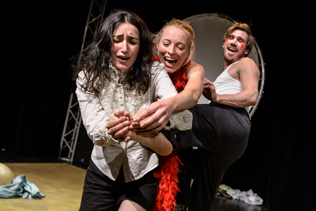 Drei Personen in Bühnenkleidung stehen eng beieinander, die vordere Person trägt ein weißes Hemd, die mittlere einen roten Federboa, die hintere ein weißes Tanktop.