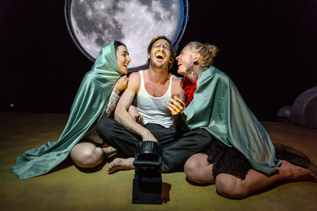 Drei Personen sitzen auf dem Boden, zwei mit grünen Umhängen umarmen eine in weißem Tanktop und schwarzer Hose, im Hintergrund ein großer runder Mond.