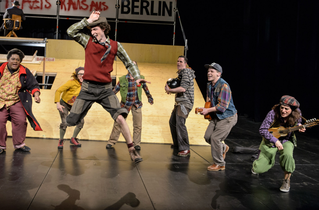 Szenenbild mit Darstellern in bunten Kostümen auf der Bühne. Einer der Schauspieler springt durch die Luft.