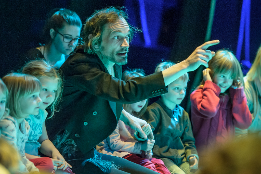 Ein Mann zeigt mit ausgestrecktem Arm nach rechts, umgeben von mehreren Kindern, die auf einer Bühne sitzen.