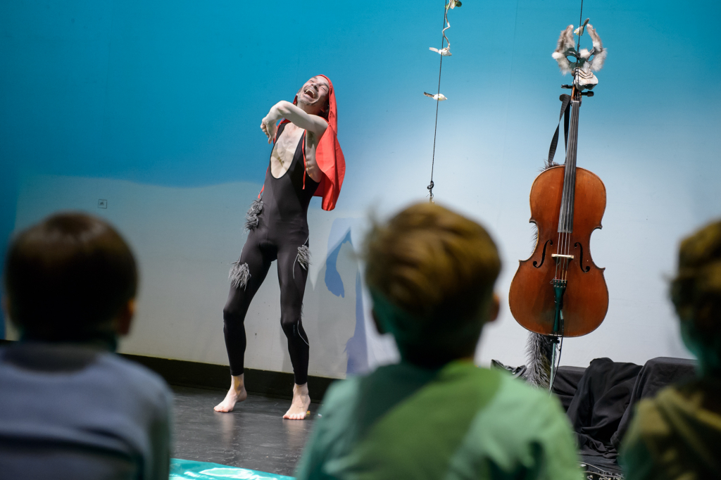 Person in schwarzem eng anliegendem Outfit und rotem Tuch auf dem Kopf tanzt vor einem Cello, mehrere Kinder schauen zu.