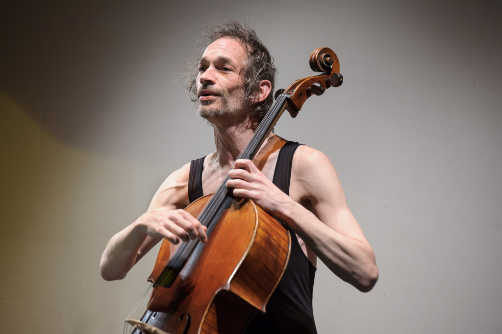 Mann mit schwarzem ärmellosem Oberteil spielt Cello, hält Bogen und Griffbrett fest.