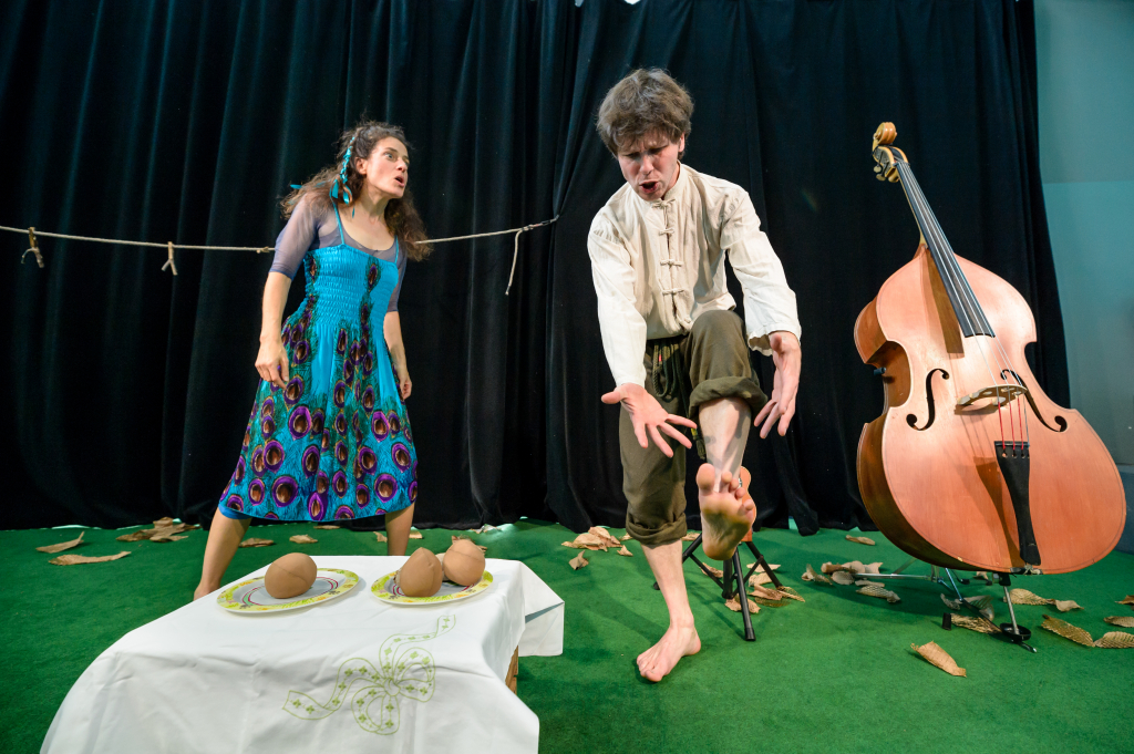 Zwei Personen in bunter Kleidung, eine hebt einen Fuß, daneben ein Kontrabass und ein Tisch mit Tellern und braunen Gegenständen.