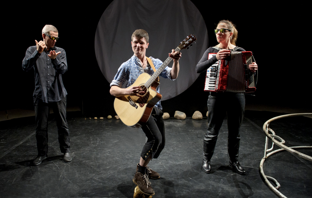 Drei Personen auf Bühne: eine spielt Gitarre, eine Akkordeon, eine gestikuliert mit Händen, im Hintergrund runde Lichtprojektion und Steine auf dem Boden.