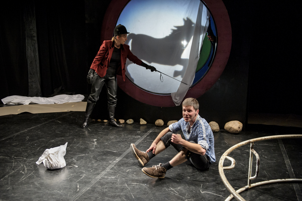 Person in rotem Jackett und schwarzer Hose steht mit Gerte vor rundem Fenster mit Schatten eines Pferdes, andere Person sitzt auf Boden mit hochgekrempelten Hosen und braunen Schuhen.