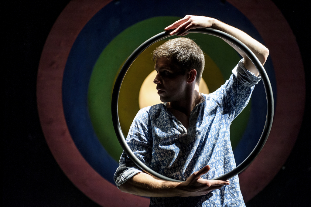 Mann mit gemustertem Hemd hält einen großen Metallring vor sich, im Hintergrund bunte konzentrische Kreise.