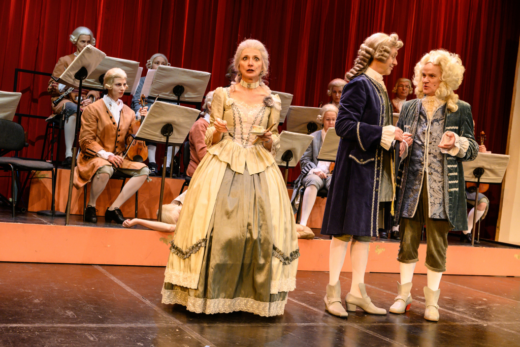 Mehrere Personen in barocken Kostümen auf einer Bühne vor roten Vorhängen, darunter eine Frau in einem cremefarbenen Kleid und zwei Männer in blauen und grauen Anzügen.