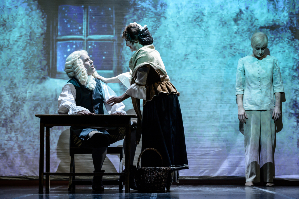 Eine Frau in historischer Kleidung steht vor einem sitzenden Mann mit Perücke und schwarzer Weste an einem Holztisch, im Hintergrund eine stehende Person in heller Kleidung.