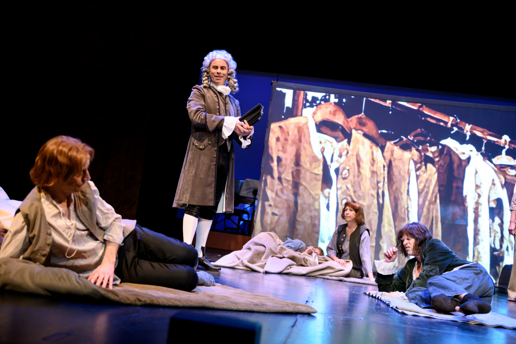 Vier Personen in historischen Kostümen auf einer Bühne, im Hintergrund eine Projektion mit mehreren Kleidungsstücken an einer Stange.