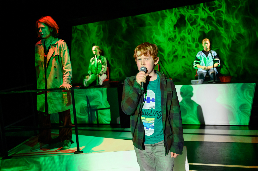 Vier Schauspieler*innen: Zwei stehen im Hintergrund, links im Vordergrund steht eine Frau in einem Manten und rechts im Vordergrund steht ein Junge, der in ein Mikrofon spricht. Grüne Lichteffekte im Hintergrund.