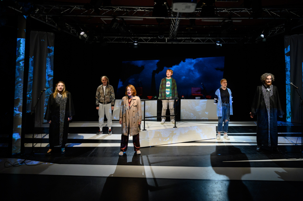 Schauspieler*innen in Roben auf der Bühne, eine Frau im Mantel als zentrale Figur im Fokus, dramatische Bühnenbeleuchtung.