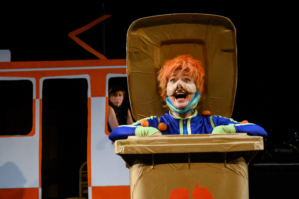 Schauspielerin in einem Müllcontainer mit orangenen Haaren und Schminke, lacht ins Publikum während einer Aufführung.