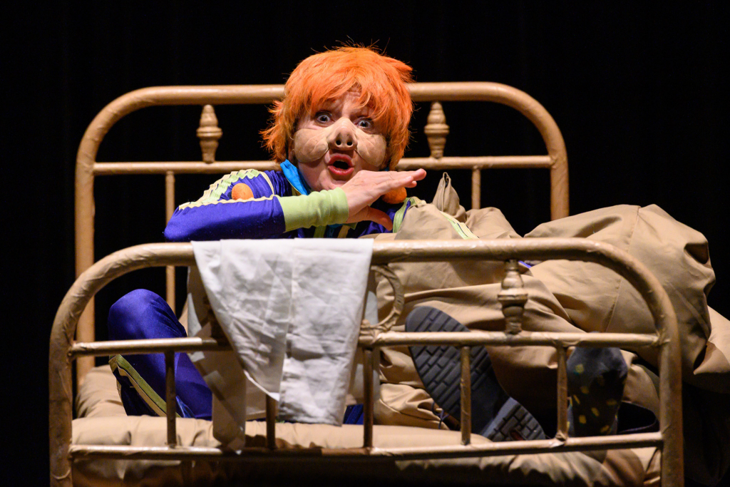 Schauspieler in einer verrückten Verkleidung auf einem Bett, mit orangefarbigem Haar und Masken.