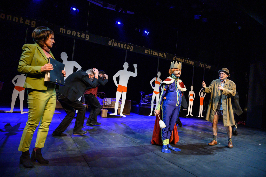 Bühne mit Schauspieler*innen in Kostümen bei einer Theateraufführung, verschiedene Charaktere interagieren.