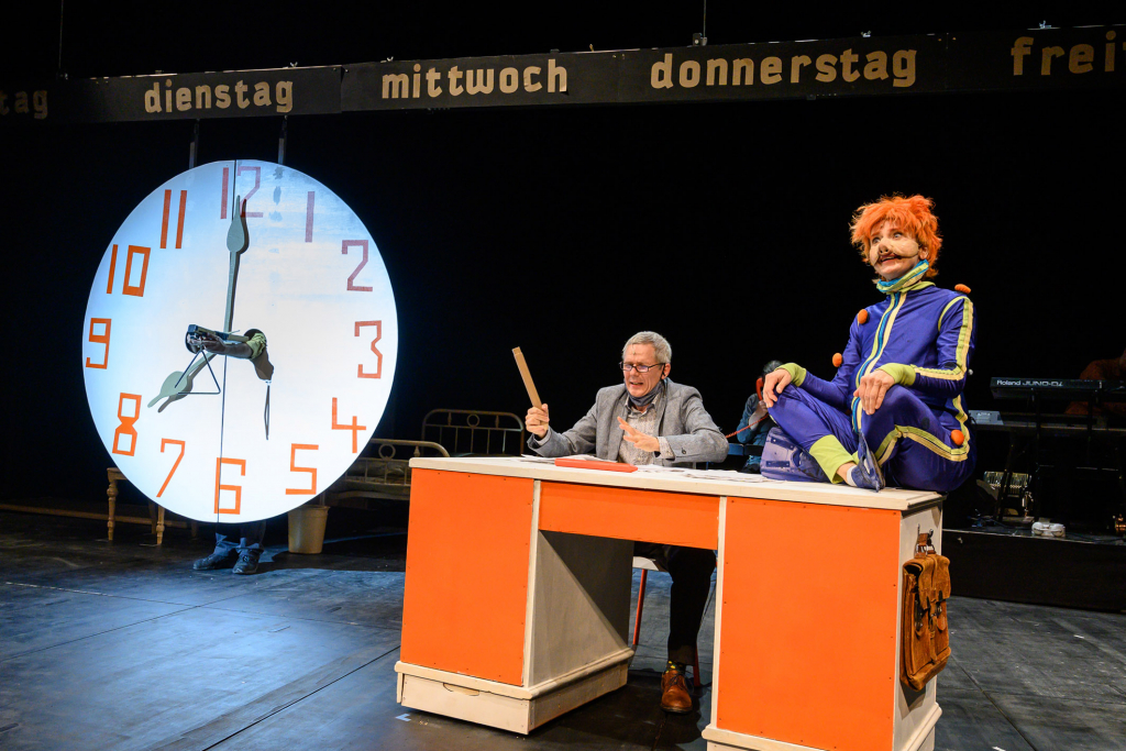 Theateraufführung mit einer großen Uhr, einem älteren Mann an einem Schreibtisch. Auf dem Schreibtisch sitzt eine weitere Person in lustigem Kostüm im Schneidersitz.
