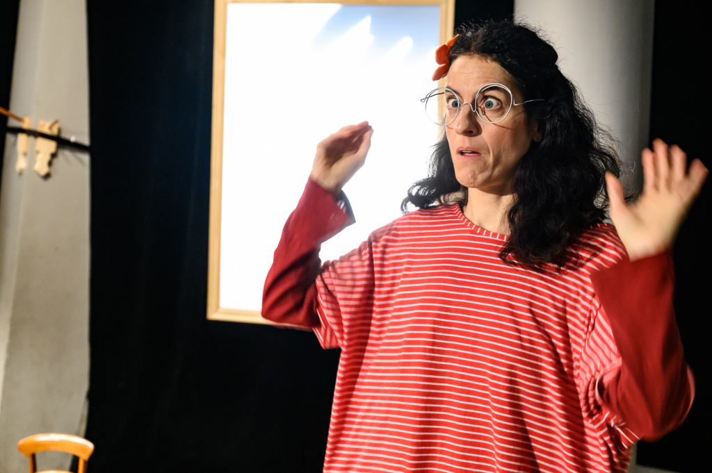 Eine Person mit Brille und lockigen Haaren steht vor einem hellen Hintergrund und hebt beide Hände, als würde sie etwas erklären oder darstellen.