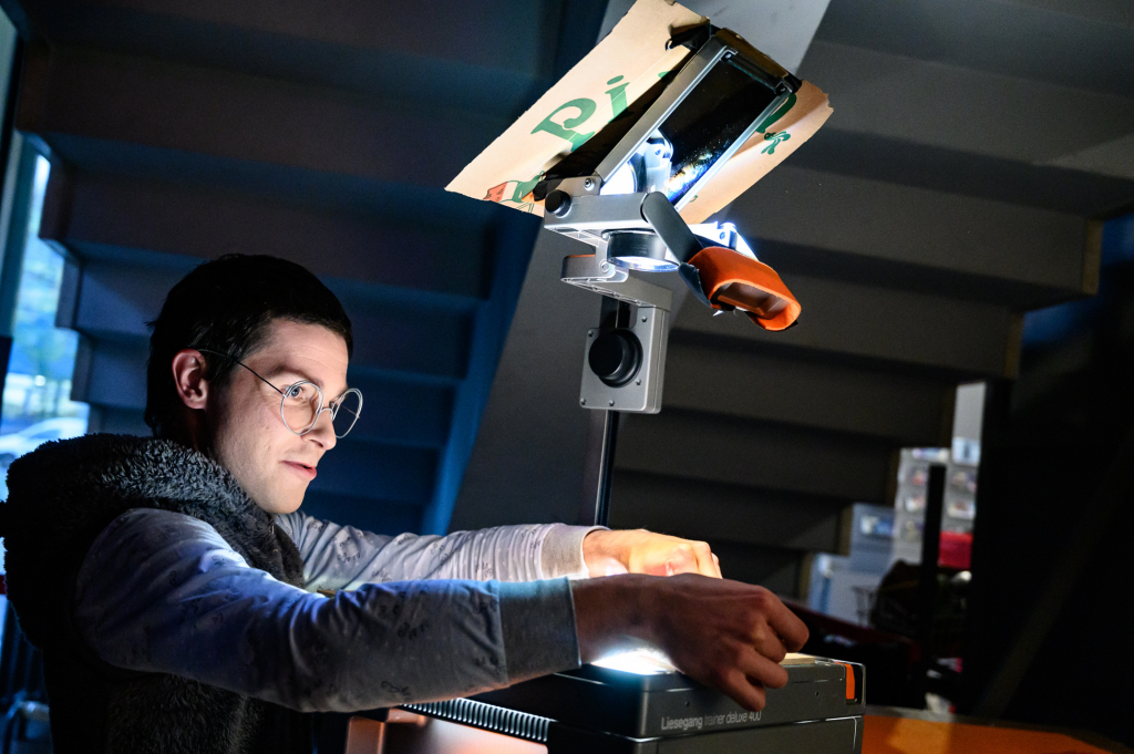 Eine Person mit Brille arbeitet konzentriert an einem beleuchteten Projektionsgerät in einem abgedunkelten Raum.