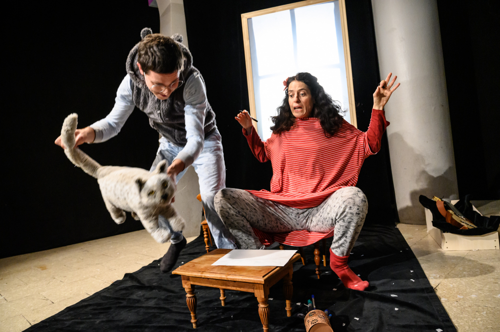 Eine Frau sitzt erschrocken an einem kleinen Tisch. Der Mann neben ihr hält eine Stoffkatze.