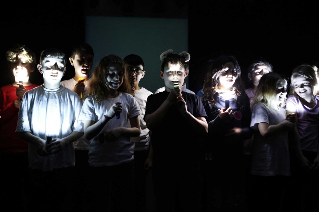Kinder auf der Bühne, die ihre Gesichter nur mit Taschenlampen beleuchten