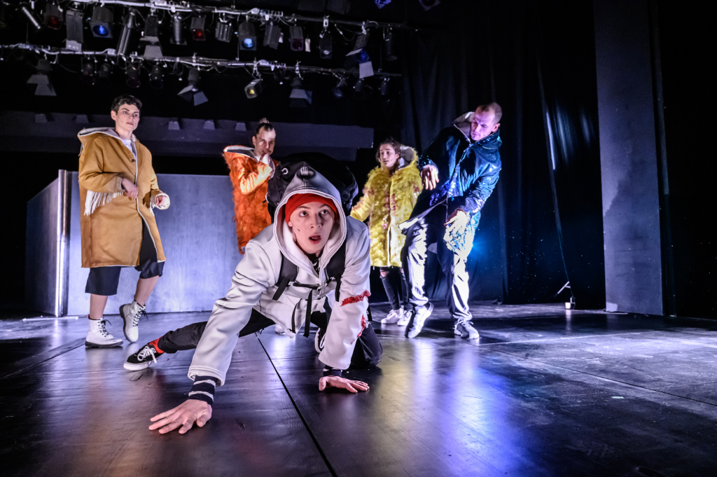 Fünf Personen auf einer Bühne, eine kniet vorn mit ausgestreckter Hand, die anderen stehen im Hintergrund in bunten Jacken.