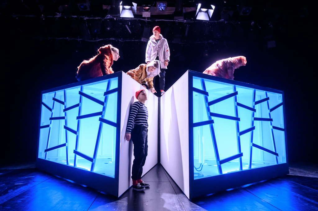Vier Personen in bunter Kleidung auf und neben zwei beleuchteten, weißen Kuben mit blauen, abstrakten Linienmustern auf einer dunklen Bühne.
