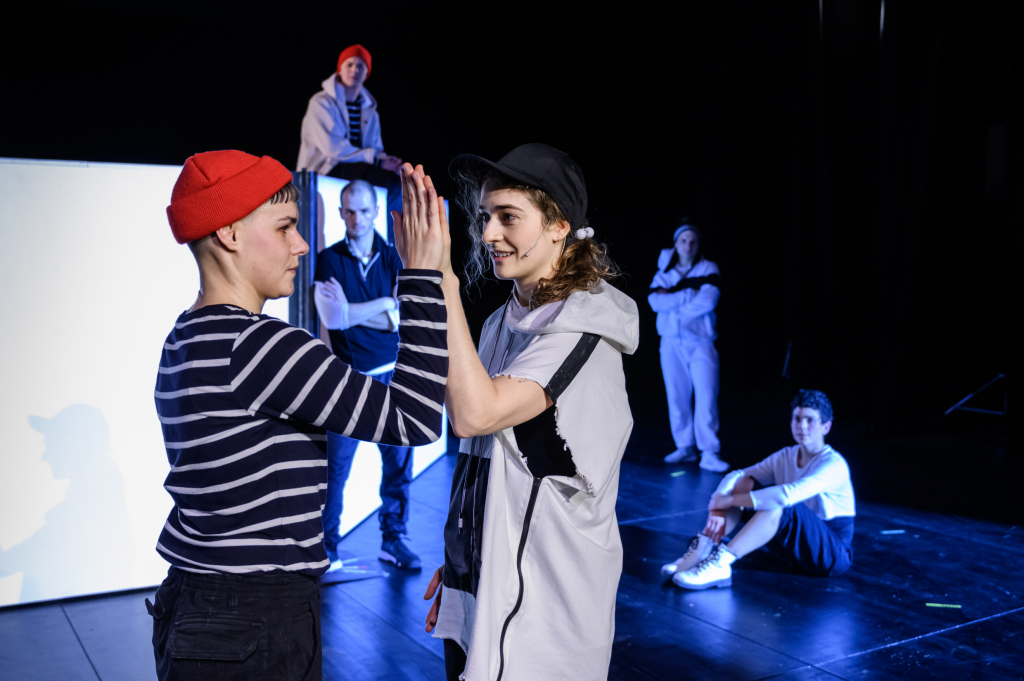 Zwei Personen stehen sich gegenüber und berühren mit erhobenen Händen, im Hintergrund weitere Personen in lockerer Haltung auf einer Bühne.