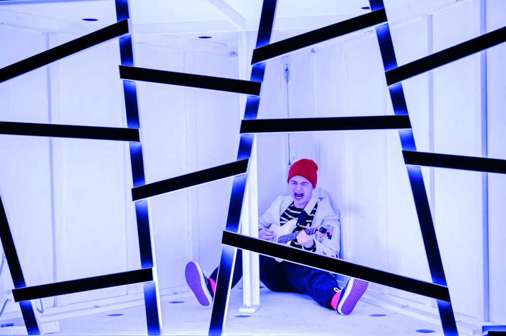 Person mit roter Mütze und weißer Jacke sitzt auf dem Boden in einem Raum mit weißen Wänden und schwarzen, schräg verlaufenden Streifen.