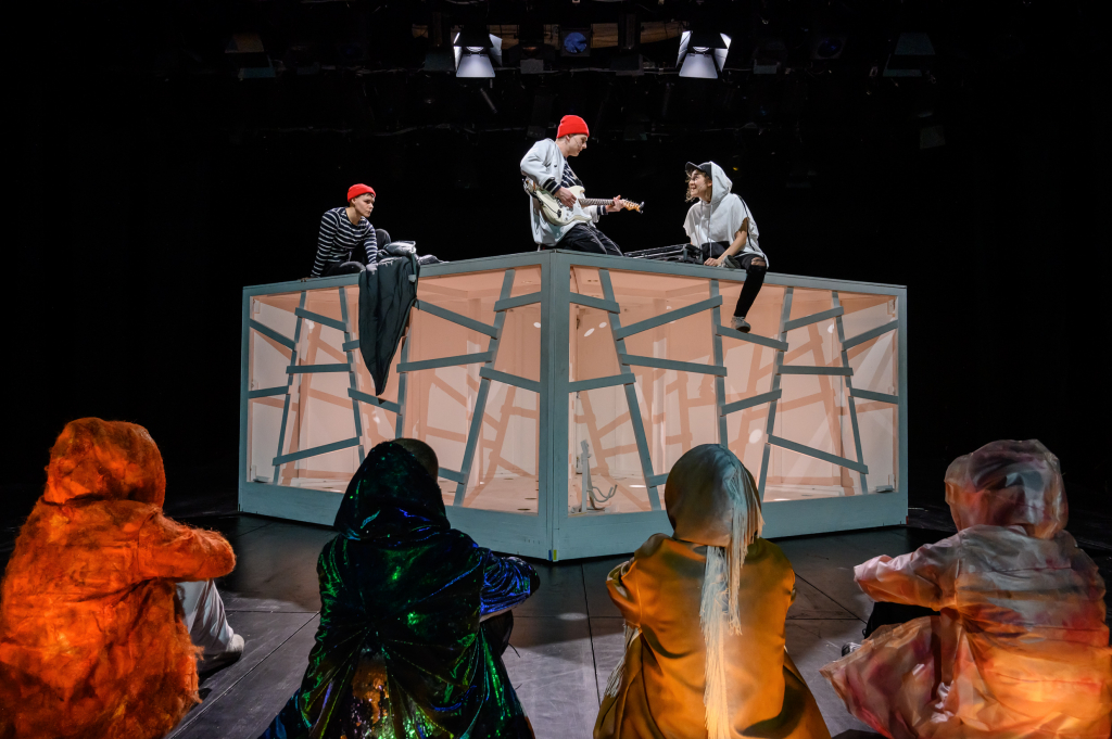 Sechs Personen in bunten, glänzenden Kostümen sitzen vor einer großen, beleuchteten, transparenten Kubusstruktur, auf der drei weitere Personen mit roten Mützen sitzen, eine spielt Gitarre.