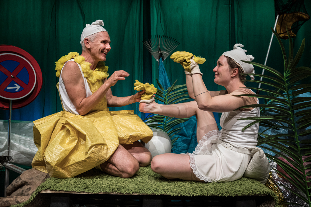 Zwei Personen in gelben und weißen Entenkostümen mit gelben Handschuhen sitzen auf einer grünen Fläche vor grünem Vorhang und Pflanzen.