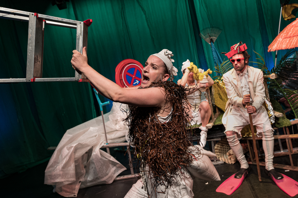 Person mit braunem, buschigem Kostüm hält eine Metallleiter, weitere Personen in weißen Kostümen mit Flossen und gelbem Entchen im Hintergrund, bunte Requisiten und grüner Vorhang.