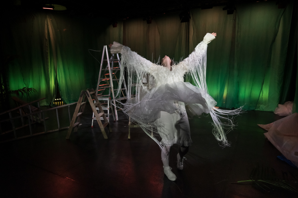 Person in weißem, fransigem Kostüm mit ausgebreiteten Armen auf dunkler Bühne mit grünem Hintergrund und zwei Leitern.