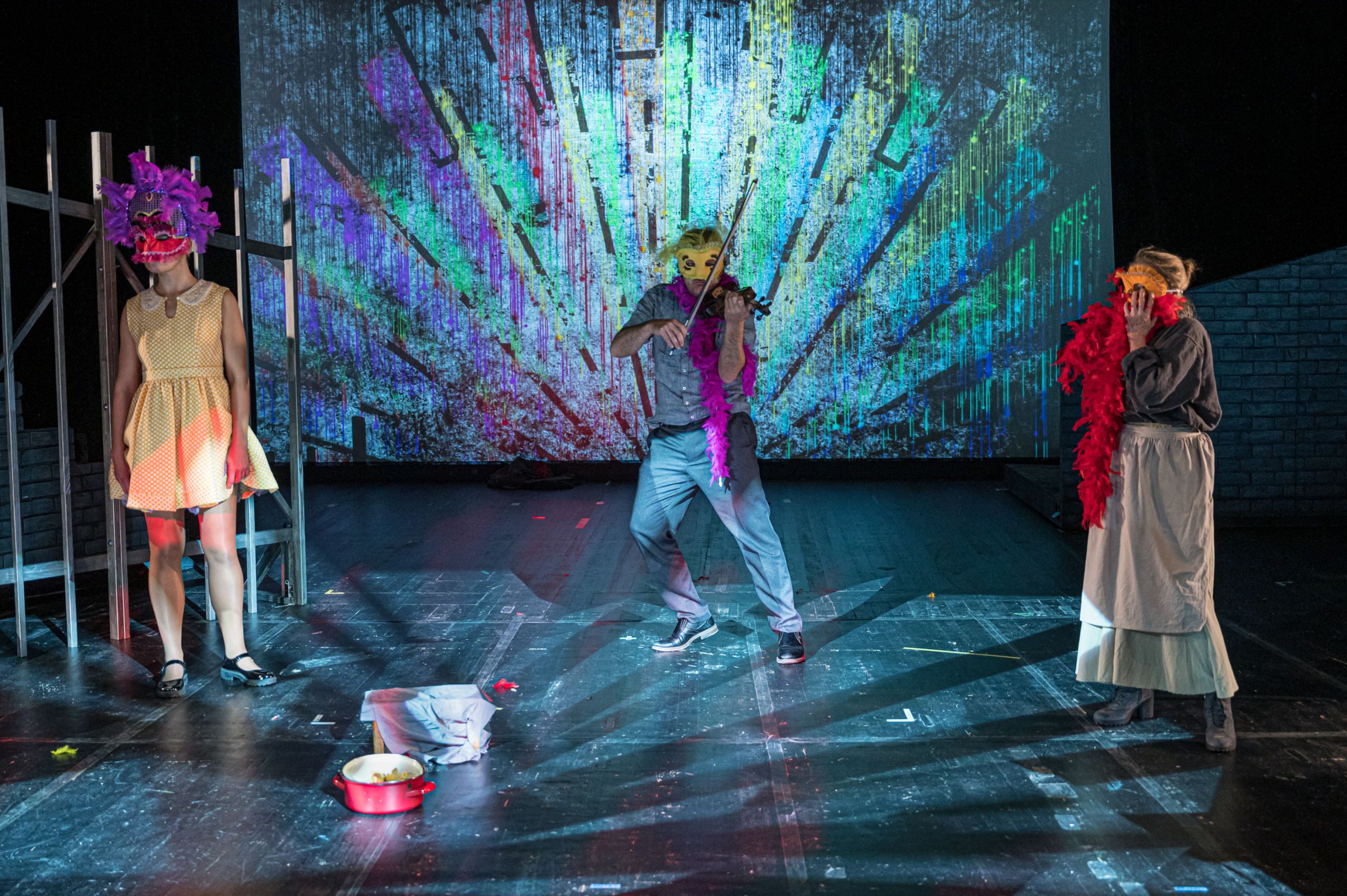 Theateraufführung mit drei Schauspieler*innen in bunten Masken und Kostümen auf einer Bühne mit kreativer Hintergrundprojektion.