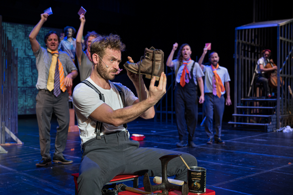 Szenenbild aus dem Theaterstück, ein Schauspieler hält einen Stiefel in der Hand während andere Darsteller im Hintergrund agieren.
