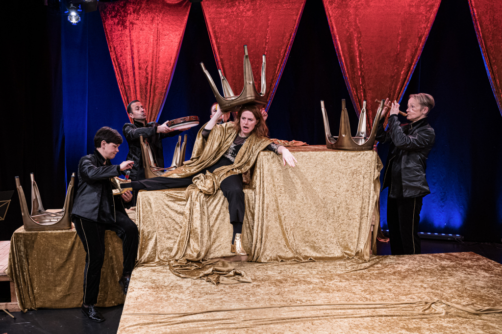 Bühnenbild des Theaters mit Schauspielern in Kostümen, glänzendem goldenen Tuch und Stühlen auf dem Tisch.