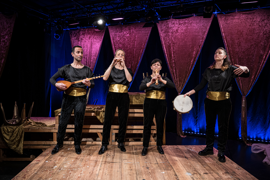 Szenenbild mit vier Musikern auf der Bühne, die verschiedene Instrumente spielen und in schwarze Kleidung mit goldenen Gürteln gekleidet sind.