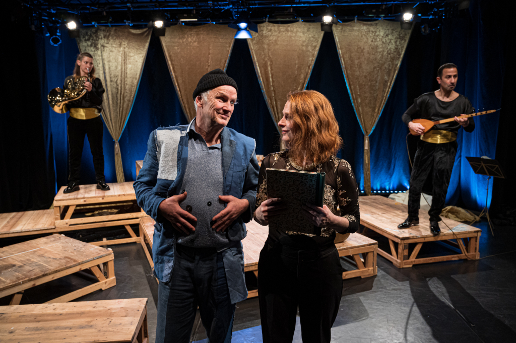 Szenenbild aus einem Theaterstück mit Schauspielern auf der Bühne in Kostümen und mit Musikinstrumenten.