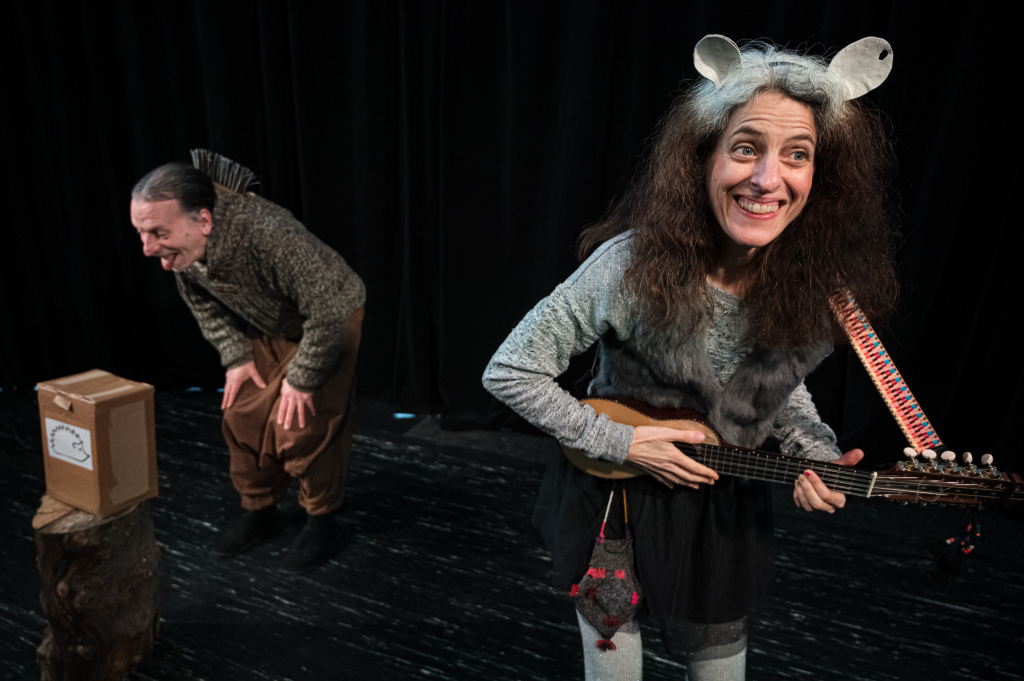 Zwei Personen auf einer Bühne, eine spielt ein Saiteninstrument, die andere beugt sich vor einem Karton mit Wolkenzeichnung auf einem Baumstumpf vor.