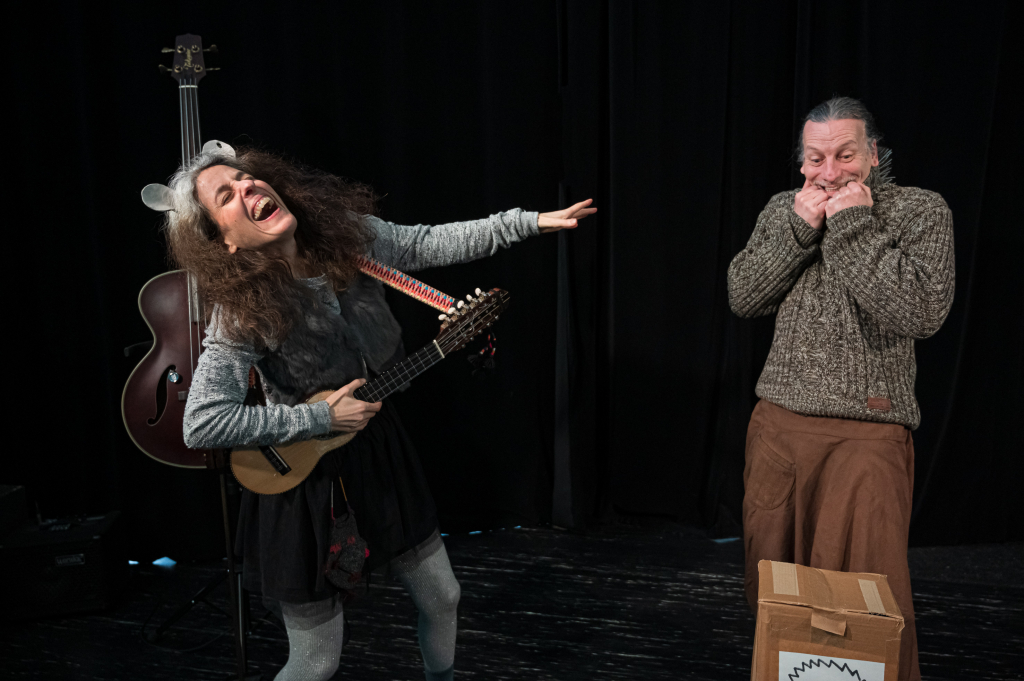 Eine Person mit langem, lockigem Haar spielt eine Ukulele und streckt den linken Arm aus, eine andere Person hält sich die Hände vor das Gesicht, beide tragen Pullover und stehen auf einer Bühne mit schwarzem Vorhang.