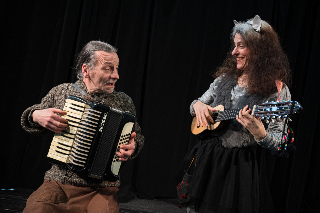 Mann spielt Akkordeon, Frau spielt Ukulele, beide vor schwarzem Hintergrund, Frau trägt Katzenohren und schwarzen Rock.